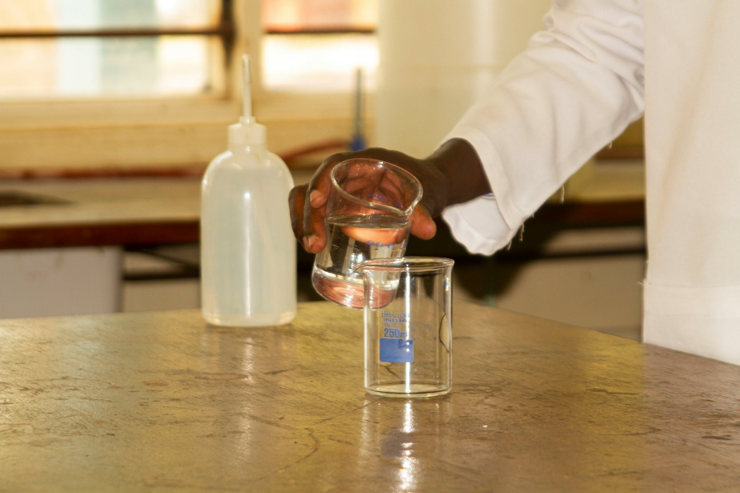 Scientist in lab coat pours liquid into beaker.